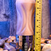 A close-up photo shows an XR Brands Tight Lipped Vibrating Vanilla Stroker with Suction + Free Movie Download standing upright on a textured surface with a yellow measuring tape beside it. The device has a flesh-toned, ribbed exterior and a metallic base featuring several control buttons for various vibrating speeds. Pebbles are scattered around the base.