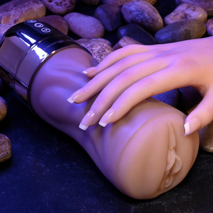A hand with neatly manicured nails is resting on a flesh-colored, cylindrical object that resembles a sex toy. The Tight Lipped Vibrating Vanilla Stroker with Suction + Free Movie Download by XR Brands, placed on a dark surface with small stones in the background, features sucking functions and vibrating speeds. The scene is softly lit.