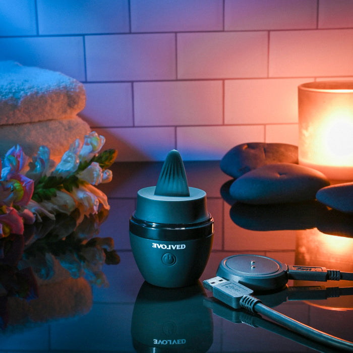 The Tongue Tied Licking Tongue Rim Job Vibrator, a rechargeable black massager, is displayed on a reflective surface with its charging cable, stones, a lit candle, flowers, and towels set against tiled decor.