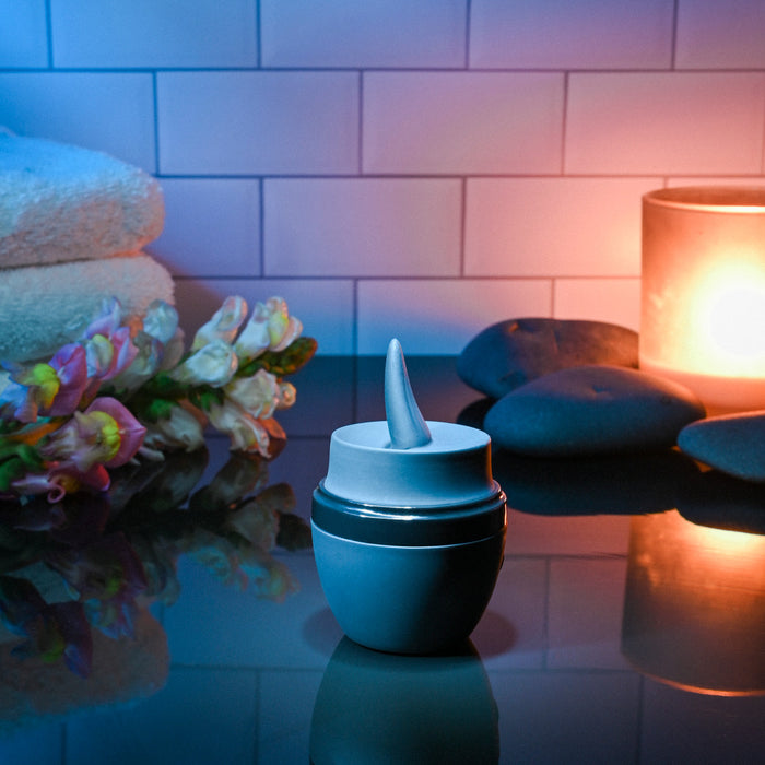 A skincare cream jar with an applicator sits on a glossy surface amid spa items and the Tongue Tied Licking Tongue Rim Job Vibrator, all arranged against a tiled wall with soft lighting for a relaxing atmosphere.