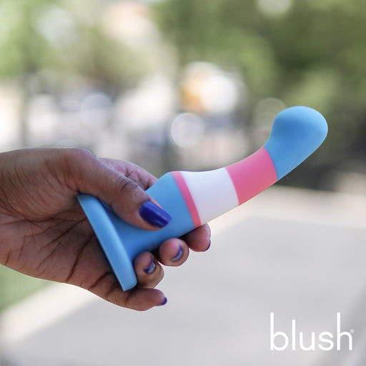 A hand with purple nails holds a colorful, curved Trans Pride 6 Inch Silicone Suction Cup Dildo, designed with blue, white, and pink stripes resembling the trans flag. Made of platinum cured silicone, this Blush toy features a suction base for versatility and includes a stimulation bump. The blurred outdoor background suggests the photo is taken outside. The brand name "Blush" is visible in the corner.