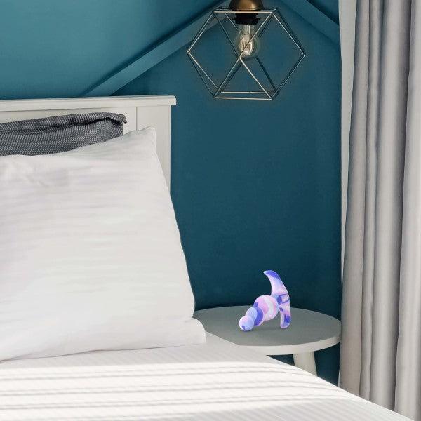 A modern bedroom features a white bed with a pillow against a teal wall and geometric pendant light. A white side table by the bed holds the vibrant Twilight Beginner Purple and Pink Silicone Butt Plug by Blush under the light, hinting at hidden pleasures for anal beginners. Tall white curtains frame the scene.