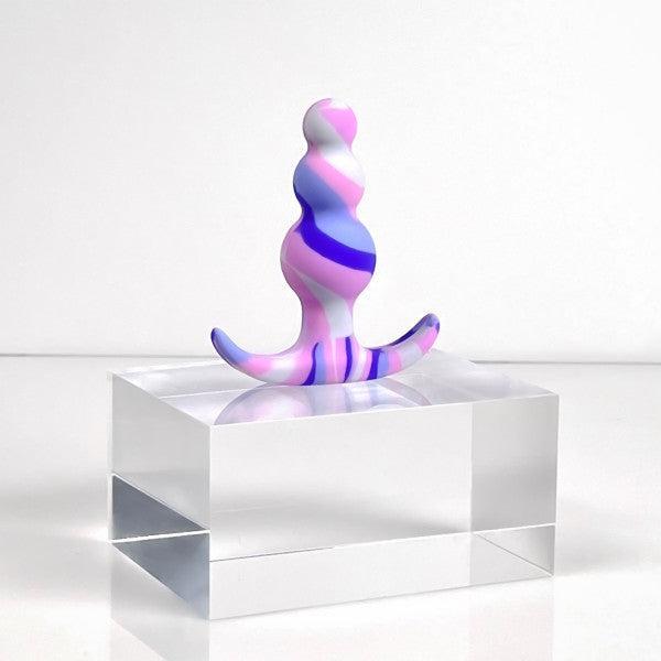 The Twilight Beginner Purple and Pink Silicone Butt Plug by Blush, with its small, twisting, curved design, stands elegantly on a clear rectangular pedestal. Crafted from platinum-cured silicone, it features vibrant swirls of pink and purple hues that create a dynamic appearance against the plain white background.