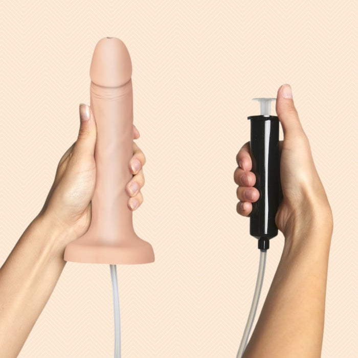 A hand holds the Strap-On-Me Realistic 7.5 Inch Silicone Squirting Dildo - Vanilla with an attached tube, while another hand holds a black handheld pump connected by the tube, against a light peach background.