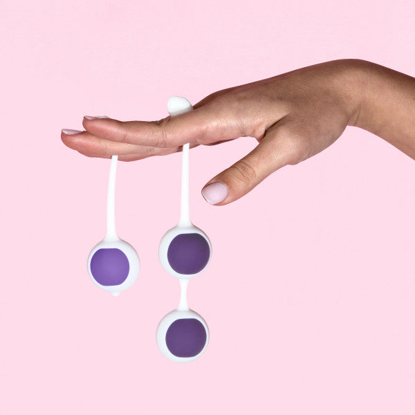 A hand with manicured nails holds two Wellness Kegel Ball Training Kit balls by their loops against a light pink background, ideal for pelvic floor strengthening.