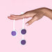 A hand with manicured nails holds two Wellness Kegel Ball Training Kit balls by their loops against a light pink background, ideal for pelvic floor strengthening.