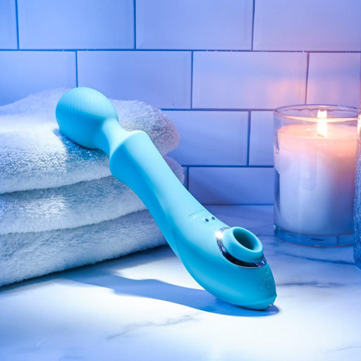 A blue silicone Wanderful Sucker Double Ended Wand + Clitoral Pulsation Vibrator by Evolved Novelties, with a curved handle and rounded head, rests on a folded white towel. Nearby, a lit candle in a glass holder casts a warm glow, enhancing the serene ambiance against the backdrop of white subway tiles.
