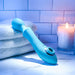 A blue silicone Wanderful Sucker Double Ended Wand + Clitoral Pulsation Vibrator by Evolved Novelties, with a curved handle and rounded head, rests on a folded white towel. Nearby, a lit candle in a glass holder casts a warm glow, enhancing the serene ambiance against the backdrop of white subway tiles.