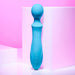 The Evolved Novelties Wanderful Sucker Double Ended Wand + Clitoral Pulsation Vibrator in blue silicone rests against abstract white geometric shapes. The background features a gradient of pink and purple tones. This handheld electric massager has three control buttons near the base.