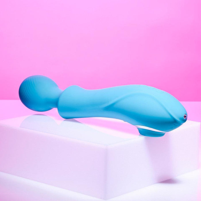 The Evolved Novelties Wanderful Sucker Double Ended Wand + Clitoral Pulsation Vibrator in blue, featuring a curved design and a textured spherical end, is displayed on a white block against a pink background. The massager showcases smooth contours and subtle button details.