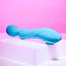 The Evolved Novelties Wanderful Sucker Double Ended Wand + Clitoral Pulsation Vibrator in blue, featuring a curved design and a textured spherical end, is displayed on a white block against a pink background. The massager showcases smooth contours and subtle button details.