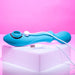 The Wanderful Sucker Double Ended Wand + Clitoral Pulsation Vibrator from Evolved Novelties, featuring a unique curved design and vibrant blue silicone finish, lies on a white surface against a pink background. The device is connected to an included white USB charging cable, which is also visible in the image.