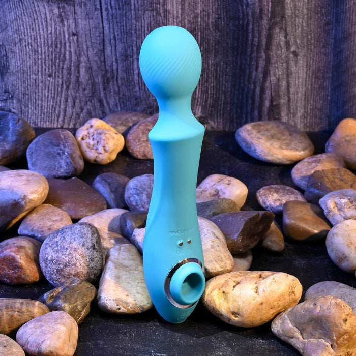 The Wanderful Sucker Double Ended Wand + Clitoral Pulsation Vibrator - Blue by Evolved Novelties rests on a bed of smooth, round stones against a dark, wooden background. This silicone massager features textured surfaces and includes a circular attachment, with a bulbous head on one end and a ring-shaped grip on the other.