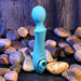 The Wanderful Sucker Double Ended Wand + Clitoral Pulsation Vibrator - Blue by Evolved Novelties rests on a bed of smooth, round stones against a dark, wooden background. This silicone massager features textured surfaces and includes a circular attachment, with a bulbous head on one end and a ring-shaped grip on the other.
