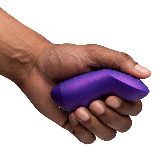 A hand holds the We-Vibe Chorus Pro Wearable Couples' Vibrator with triple motors and remote, a small, purple, curved couple’s toy for G-spot stimulation, against a white background.