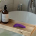 A wooden bath tray holds a We-Vibe Melt 2 Rechargeable Pleasure Air Clitoral Stimulator in purple and a brown liquid soap bottle, with a white bathtub and faucet visible in the background.