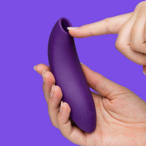 A hand holds the We-Vibe Melt 2 Rechargeable Pleasure Air Clitoral Stimulator as another hand points to its round opening, highlighting its waterproof, smooth silicone design against a solid purple background.
