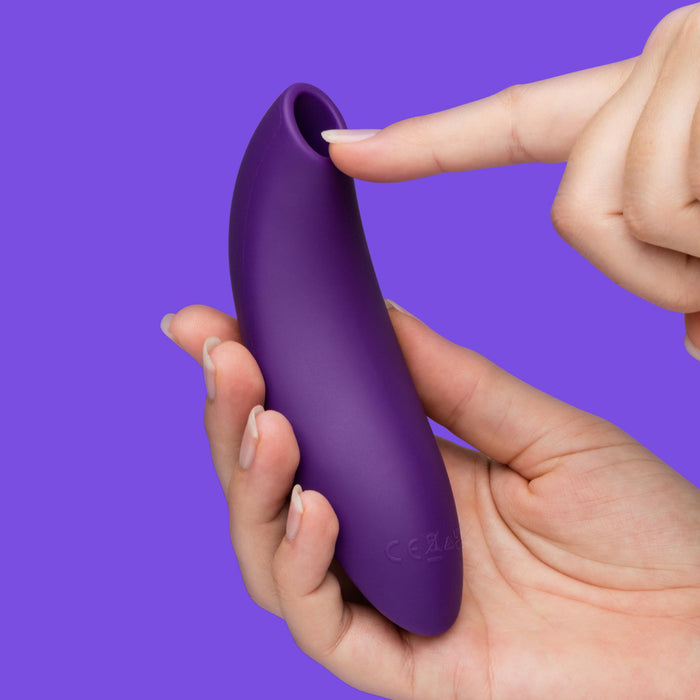 A hand holds the We-Vibe Melt 2 Rechargeable Pleasure Air Clitoral Stimulator as another hand points to its round opening, highlighting its waterproof, smooth silicone design against a solid purple background.