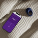 A smartphone with a purple “PIVOT” app screen and the We-Vibe Pivot Vibrating Silicone Rechargeable Penis Ring rest on a beige striped fabric, sunlight casting gentle shadows across them.
