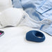 The We-Vibe Pivot Vibrating Silicone Rechargeable Penis Ring, in blue, rests on a white surface beside a white lace bra, a blue shirt, and part of a smartphone.