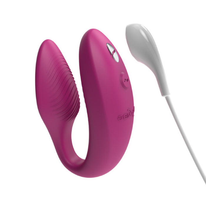 The We-Vibe Sync Remote and App Controlled Wearable Couples Vibrator in Dusty Pink, by We-Vibe, is U-shaped and designed for couples. It includes a remote control, with one end featuring a ribbed texture and the other end having a smooth surface. The product comes with a white cord that has a rounded tip connected to the remote.