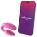 A We-Vibe Sync Remote and App Controlled Wearable Couples Vibrator in Dusty Pink with a textured surface sits next to a black smartphone displaying the "We-Vibe" logo on a purple and red gradient background.