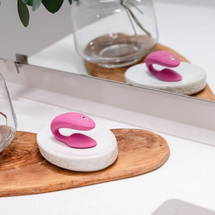 A dusty pink, curved We-Vibe Sync Remote and App Controlled Wearable Couples Vibrator with a silicone texture is displayed on a white and wooden platform next to a wall mirror. The reflection of the We-Vibe product is visible in the mirror, and a glass jar with green leaves is seen in the background.