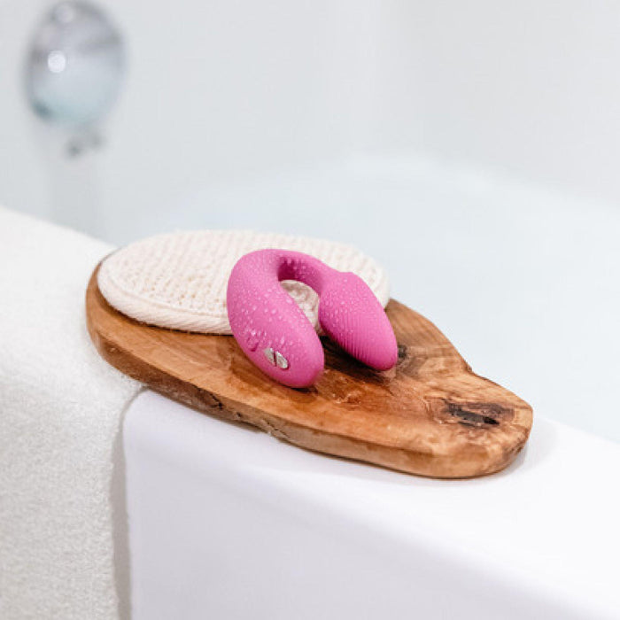 A small, dusty-pink U-shaped We-Vibe Sync Remote and App Controlled Wearable Couples Vibrator with a textured surface rests on a wooden board along with a round, beige, textured pad. The board is placed on a white bathroom edge near a filled bathtub with a blurred chrome faucet in the background.