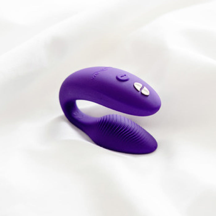 A purple, C-shaped silicone wearable couples' vibrator with dual ends and control buttons, resting on a white fabric surface. The We-Vibe Sync Remote and App Controlled Wearable Couples Vibrator - Purple by We-Vibe includes a wireless remote control for added convenience.