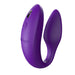 The We-Vibe Sync Remote and App Controlled Wearable Couples Vibrator in purple is designed in a C-shape with a ribbed texture on one end. This device features a small button and the brand name "We-Vibe" engraved on its surface, and it’s set against a white background.