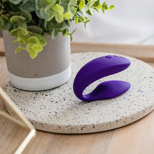 A We-Vibe Sync Remote and App Controlled Wearable Couples Vibrator in purple, featuring a curved design and textural details, is elegantly placed on a round speckled surface. In the background, a potted plant with lush green leaves adds to the modern and serene ambiance of the minimalist indoor setting.
