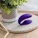 A We-Vibe Sync Remote and App Controlled Wearable Couples Vibrator in purple, featuring a curved design and textural details, is elegantly placed on a round speckled surface. In the background, a potted plant with lush green leaves adds to the modern and serene ambiance of the minimalist indoor setting.