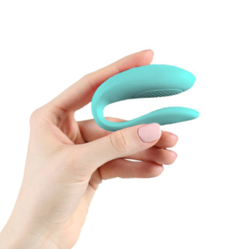 A light-skinned hand holds an aqua, U-shaped We-Vibe Sync Lite App Controlled Wearable Couples Vibrator against a plain white background, showcasing its sleek design and compatibility with the We-Vibe App.