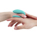 Two hands are shown holding a small, aqua, U-shaped device. The left hand, which appears to belong to a lighter-skinned adult, holds the We-Vibe Sync Lite App Controlled Wearable Couples Vibrator between their thumb and forefinger. The right hand supports the left hand from below. The background is white.