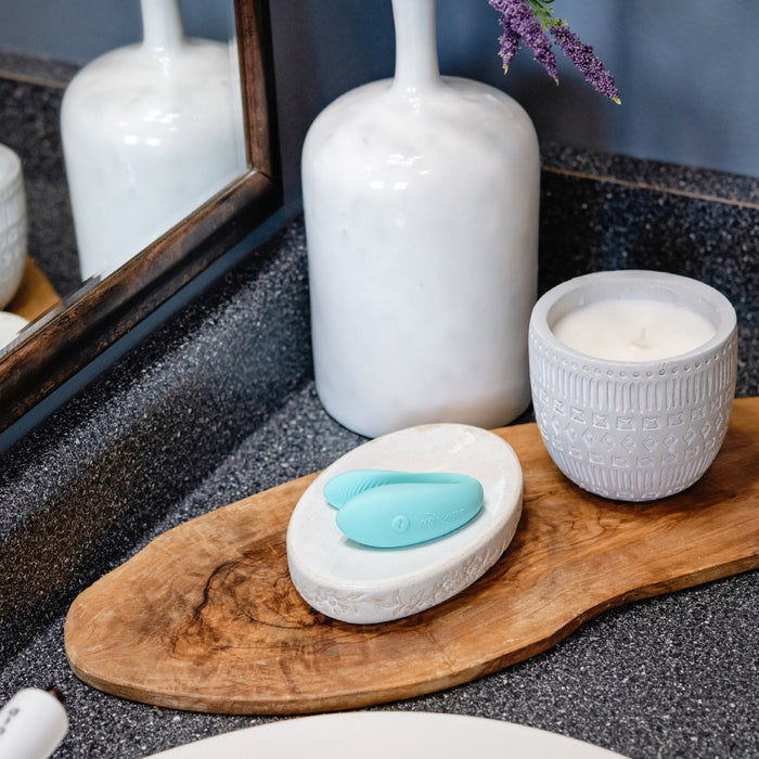A bathroom countertop with a wooden tray holds a white soap dish with a turquoise silicone facial cleansing brush on it. Next to the soap dish is a white decorative candle, a ceramic vase with a purple flower, and—tucked discreetly—a We-Vibe Sync Lite App Controlled Wearable Couples Vibrator in Aqua for an added touch of intimacy. A portion of a mirror is also visible.