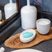 A bathroom countertop with a wooden tray holds a white soap dish with a turquoise silicone facial cleansing brush on it. Next to the soap dish is a white decorative candle, a ceramic vase with a purple flower, and—tucked discreetly—a We-Vibe Sync Lite App Controlled Wearable Couples Vibrator in Aqua for an added touch of intimacy. A portion of a mirror is also visible.