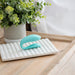 The We-Vibe Sync Lite App Controlled Wearable Couples Vibrator in Aqua is placed on a rectangular, white, ribbed tray on a wooden surface. A green potted plant positioned behind the tray adds a touch of natural decor to the scene.