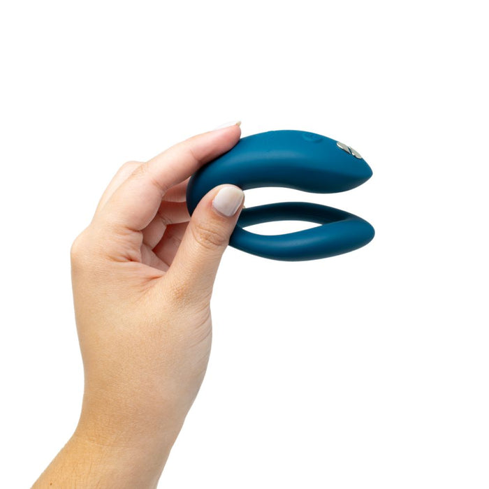 A hand is holding a We-Vibe Sync O Hands-Free Wearable Couples Vibrator in green, featuring a U-shaped design with smooth curves and a sleek aesthetic against a white background. This app-controlled vibe for couples has dual, slightly tapered ends.