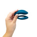 A hand is holding a We-Vibe Sync O Hands-Free Wearable Couples Vibrator in green, featuring a U-shaped design with smooth curves and a sleek aesthetic against a white background. This app-controlled vibe for couples has dual, slightly tapered ends.