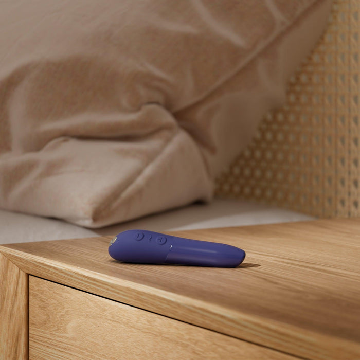 The We-Vibe Tango X Powerful Bullet Vibrator in blue rests on a wooden bedside table next to a beige bed with a rattan headboard. The table and bed are part of the cozy bedroom setup. The device stands out due to its bright color against the neutral tones of the room.