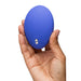 A hand holds the We-Vibe Temp Lay On Vibrator with Hot and Cold Functions, a blue oval-shaped device featuring two silver buttons, set against a plain white background.