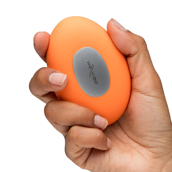 A hand holds the orange, oval-shaped We-Vibe Temp Lay On Vibrator with Hot and Cold Functions, featuring a silver "WE-VIBE" button, against a white background.