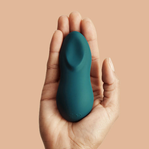 A hand holds the We-Vibe Touch X Lay On Vibrator in green, a small silicone device with a smooth, rounded shape, against a beige background.