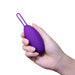 A hand is holding the Blush Wellness Imara Wearable Vibrating Egg with Remote - Purple, a small oval-shaped vibrator with a loop at one end, against a white background.