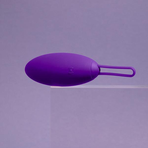 The Blush Wellness Imara Wearable Vibrating Egg with Remote - Purple, oval-shaped and equipped with a loop handle at the end, rests on a transparent surface against a light purple background. This sleek silicone device boasts 10 vibration modes and features a wireless remote control for ultimate convenience.