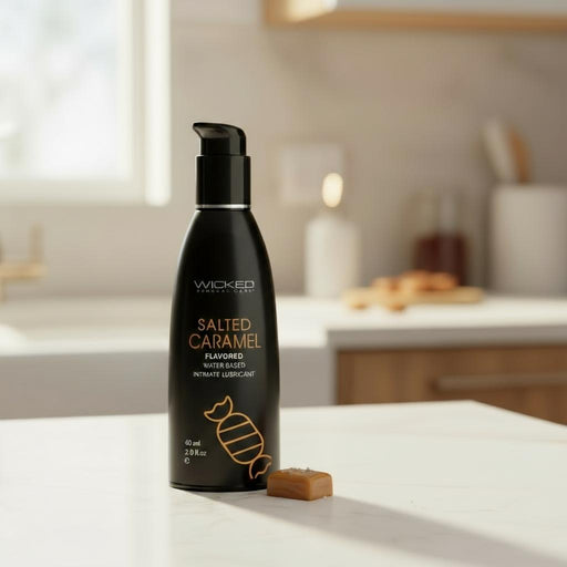 A 2oz bottle of Wicked Aqua Salted Caramel Flavored Water Based Lubricant sits on a kitchen counter next to caramel candy, with a softly lit kitchen in the background.