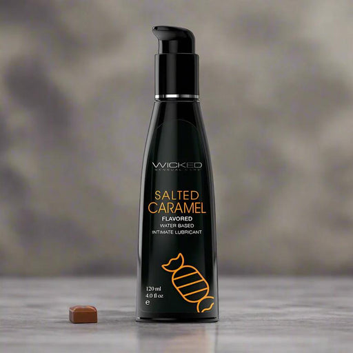 A 4 oz black bottle of Wicked Aqua Salted Caramel Flavored Water Based Lubricant stands upright on a gray surface with a caramel candy, set against a blurred gray background. This lubricant is sugar-free and deliciously smooth.