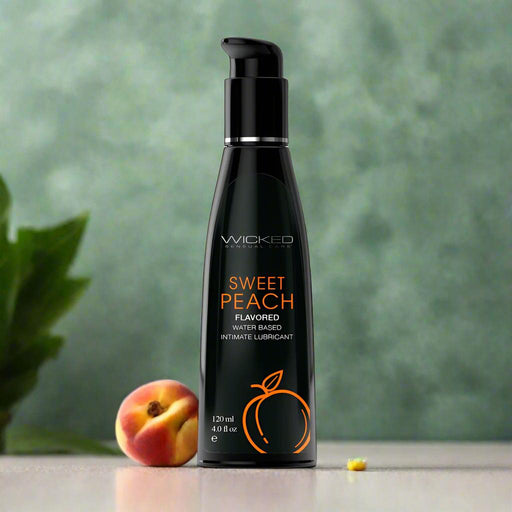 A black bottle of Wicked Aqua Sweet Peach Flavored Water Based Lubricant 4 oz sits on a smooth surface, with an orange peach outline on the label. A real peach and a yellow flower in the background accentuate its sugar-free allure.