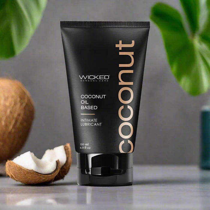 A black tube of Wicked Coconut Oil Based Lubricant - 4 oz stands upright, with coconut halves and green leaves in the background. "Coconut" is printed vertically on the tube; it’s perfect as a sensual massage oil or vulva moisturizer.
