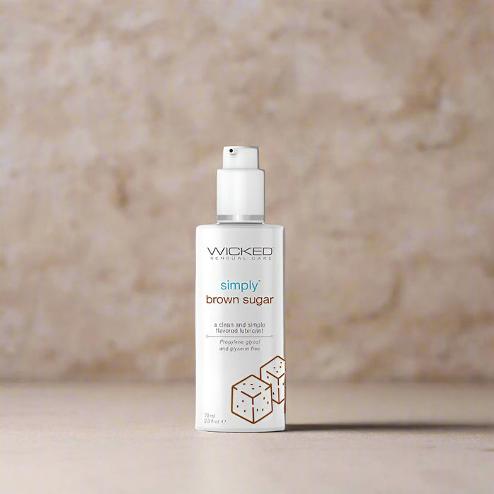 A white bottle labeled "Wicked Simply Brown Sugar Flavored Water Based Lubricant 2.3 oz" from Wicked Lubes, featuring a pump dispenser on top. The design showcases images of sugar cubes and text highlighting that it is a clean, simple, glycerin-free brown sugar flavored lube with a pH balanced formula.